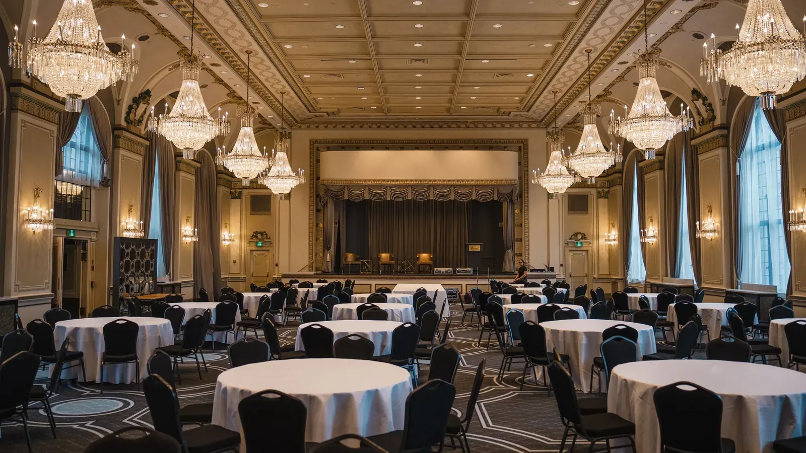 An empty ballroom with coffered ceiling and crystal chandeliers is set with round tables and black chairs facing a stage.