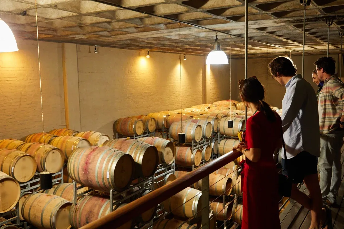 A small group stands along a wooden railing on a raised walkway, looking down at rows of oak barrels stacked on metal racks in a brick cellar. Pendant lamps hang from the coffered ceiling and cast warm light across the barrels.