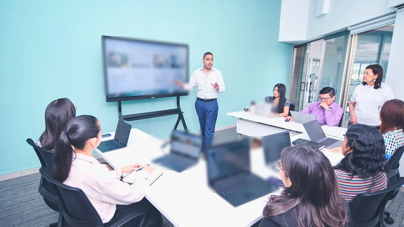 A presenter stands beside a large screen and walks a team through his material in a bright training room. Colleagues with open laptops sit around white tables and follow along.