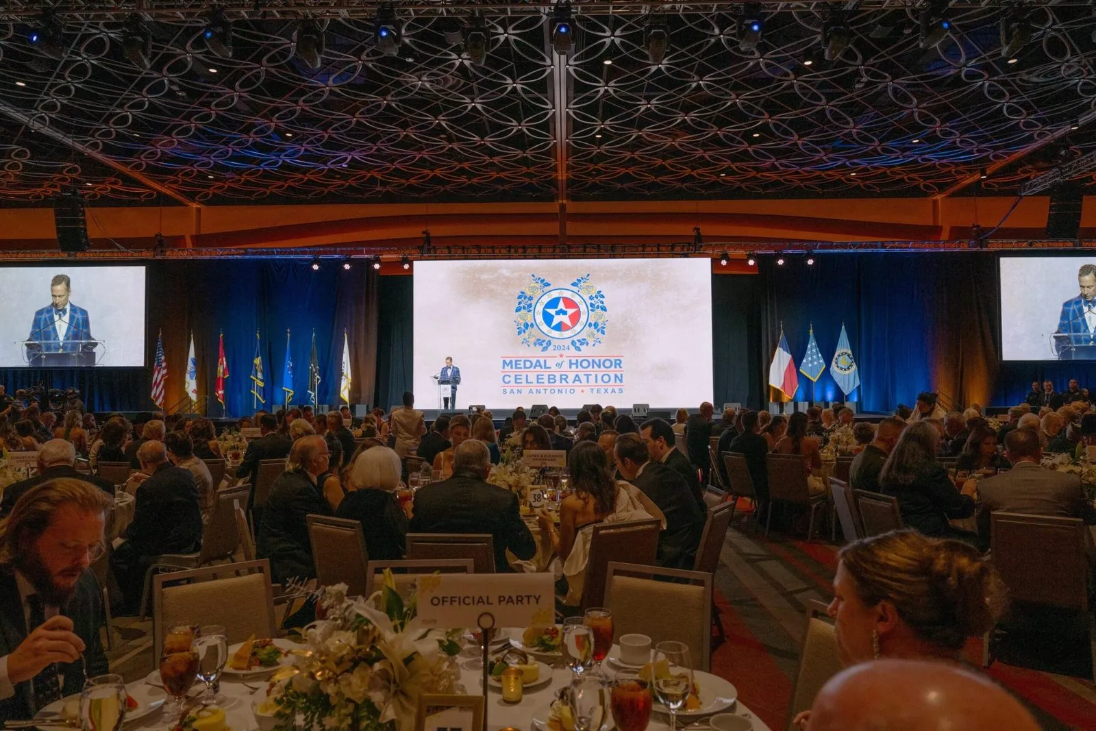 Speaker at a lectern in a banquet hall with flags and screens