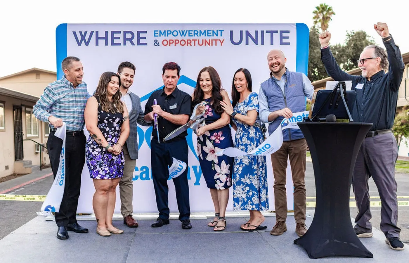 Group of people on an outdoor stage at a ribbon cutting with a banner behind them