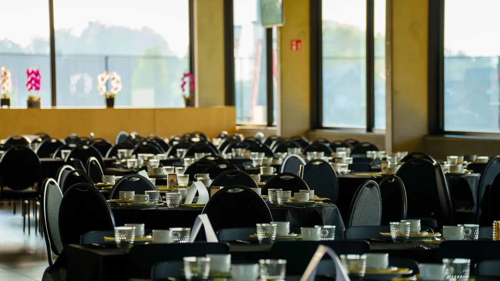 Tables with black chairs and place settings fill a bright event hall with large windows, set for a group dinner.