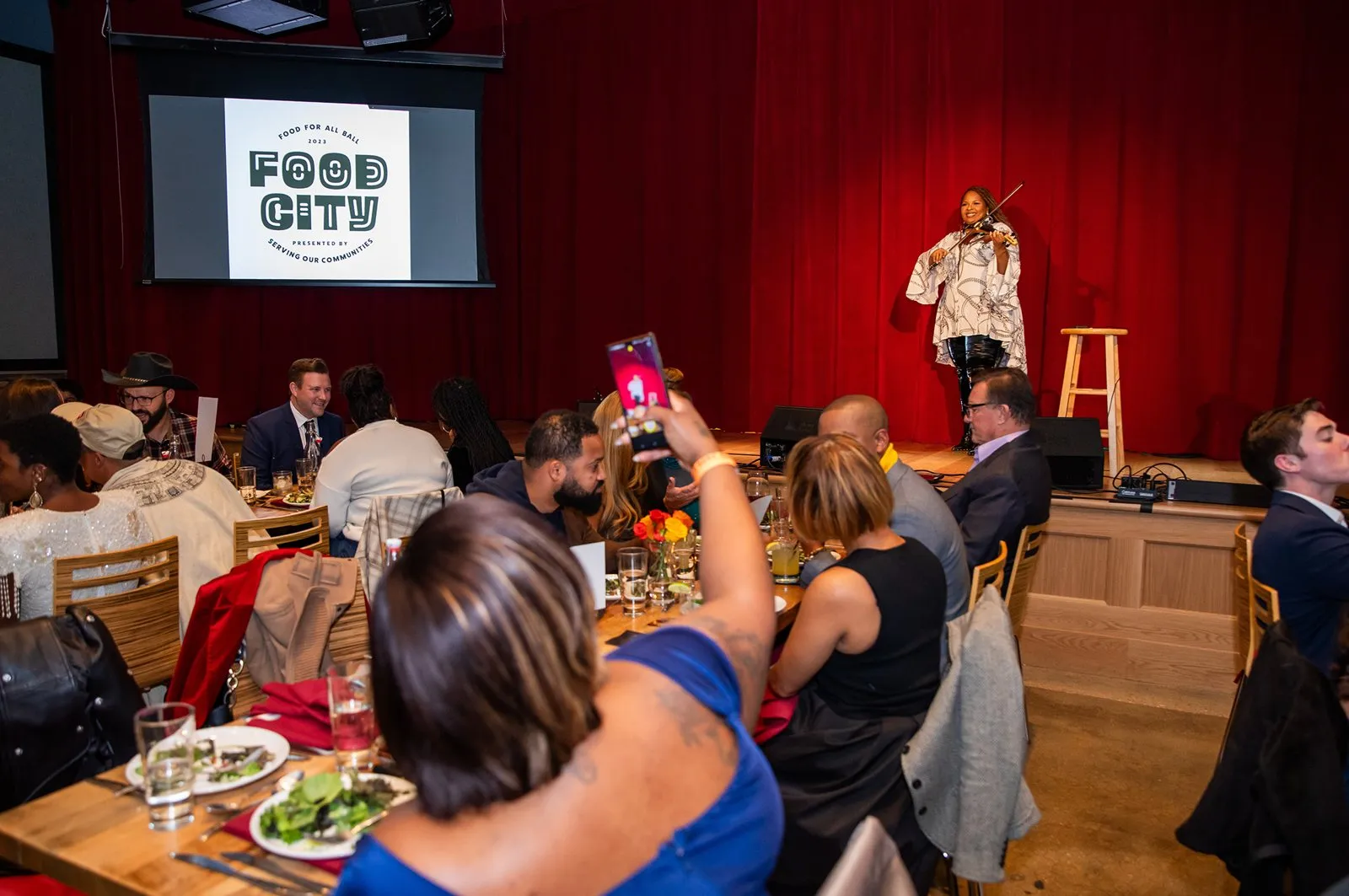 Dinner guests at long tables facing a stage with a Food City screen