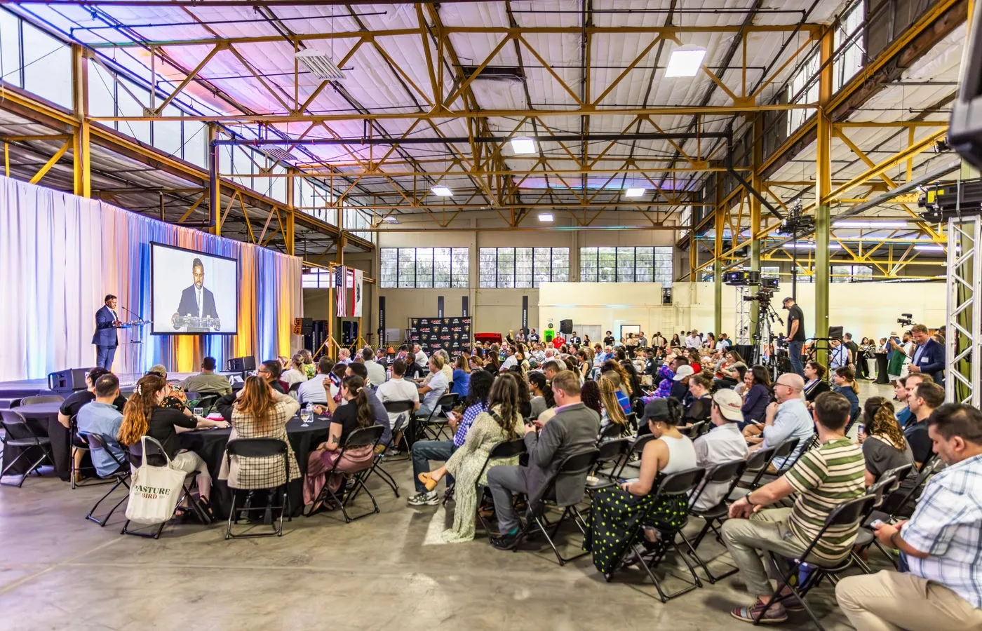 Audience seated in a large industrial hall with screens and string lights