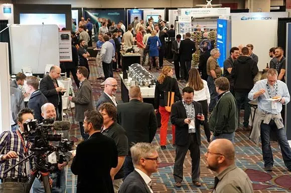 Busy exhibit hall with attendees talking and a camera crew filming