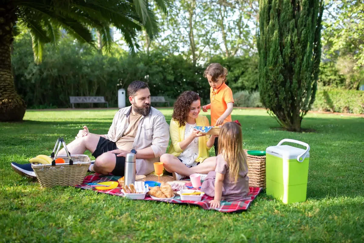 A family shares a relaxed picnic on a sunny lawn, with croissants and fruit spread on a plaid blanket. The children help pass food while the parents chat, an easy daytime gathering in the park.