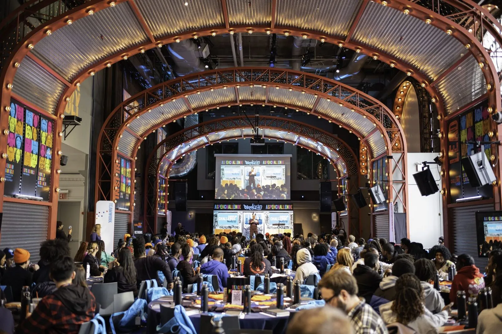 Audience seated in a theater style hall facing a stage screen