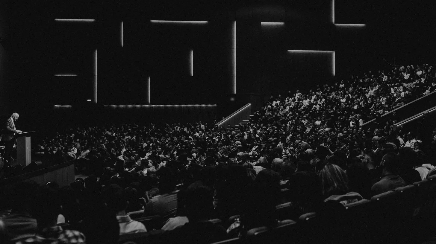 A large audience fills a dark auditorium while a speaker stands at a lectern on stage at a national sales meeting.
