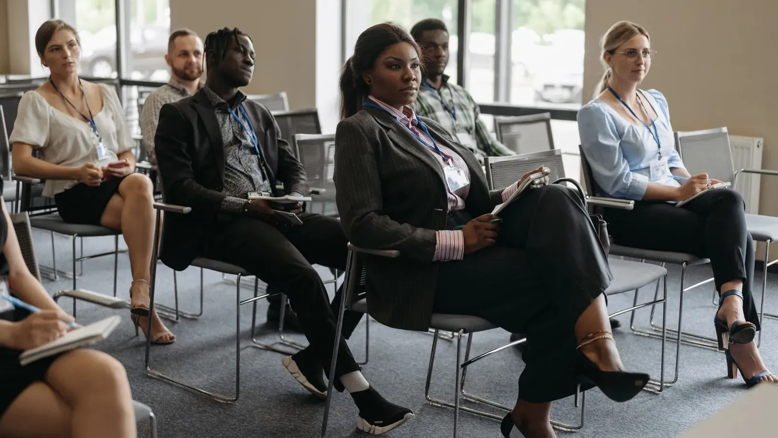 Six attendees with lanyards sit in rows of gray chairs taking notes during a session in a bright meeting room.