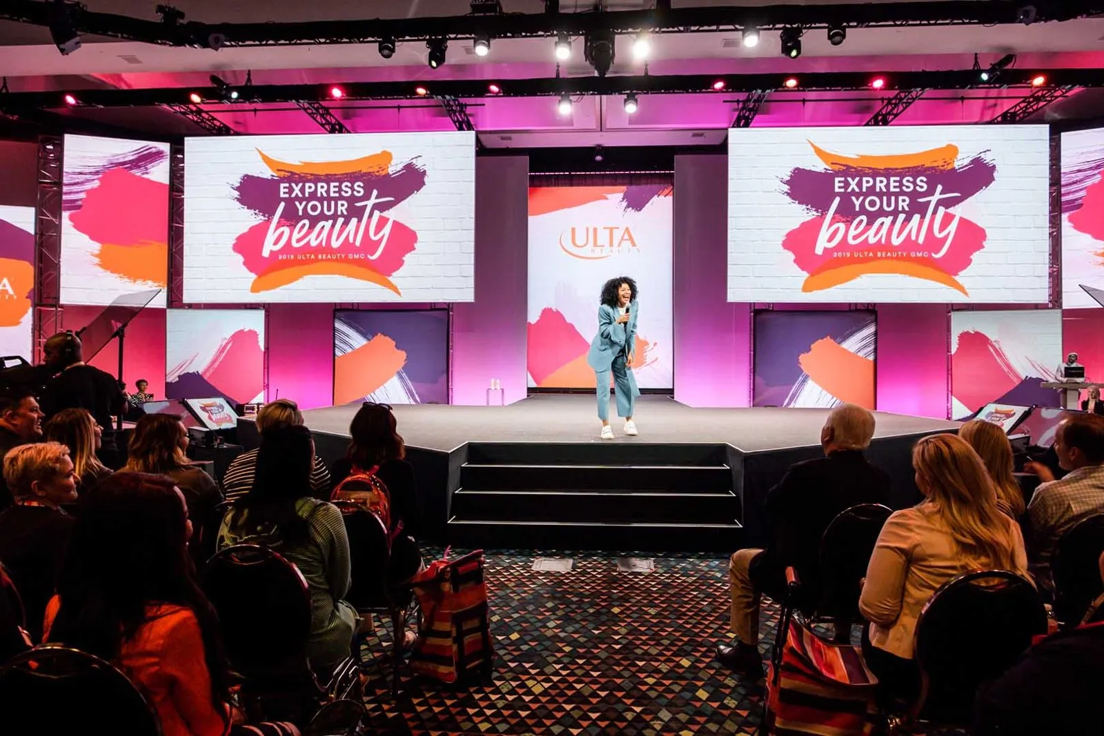 Conference stage with a presenter and large Express Your Beauty screens