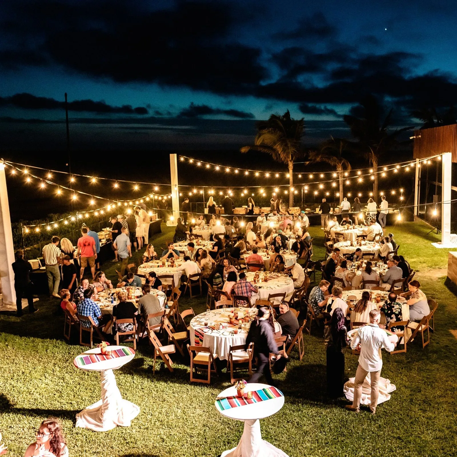 Round dinner tables on a lawn at dusk under string lights