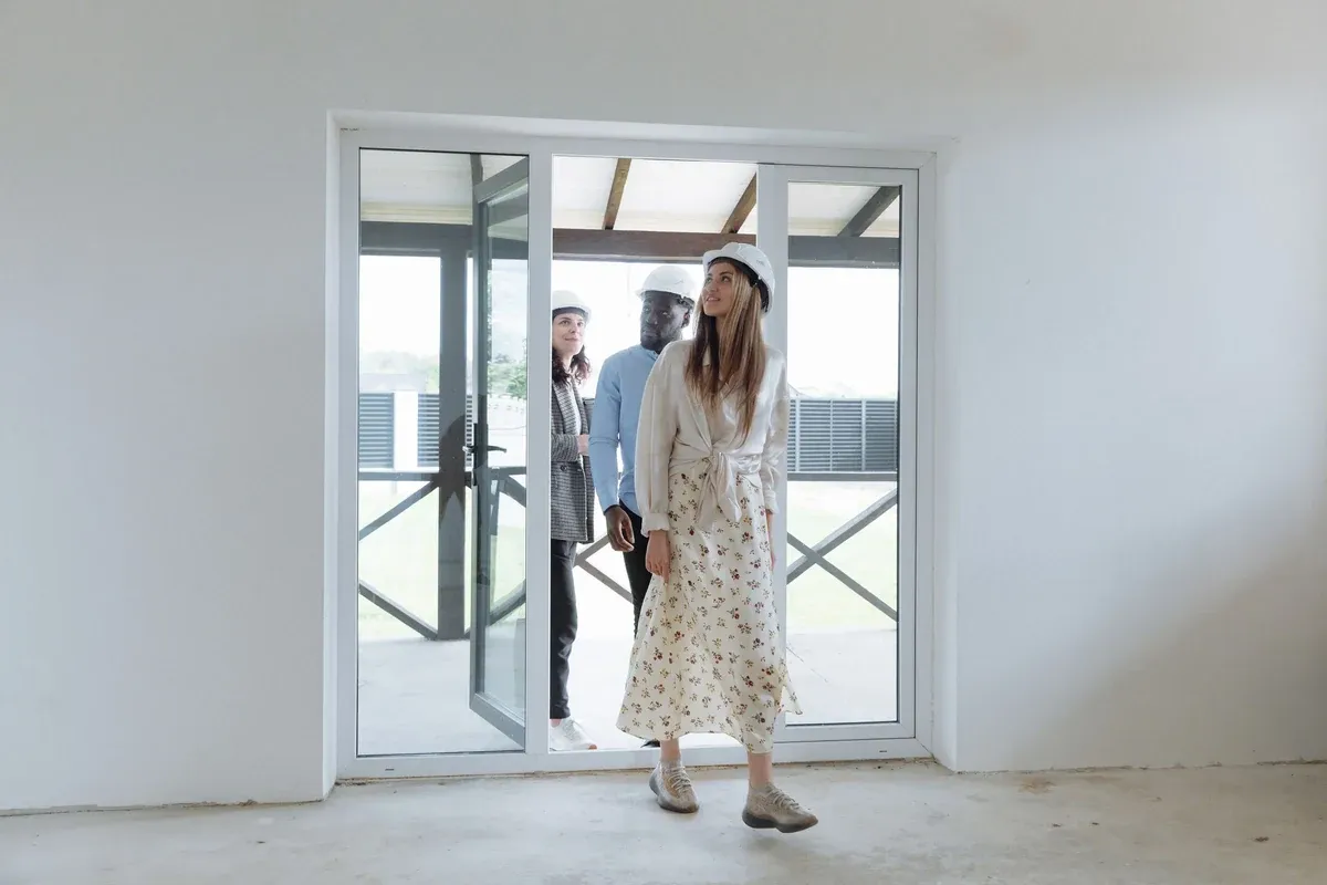 Three people in white hard hats step through glass patio doors into a bright, unfinished room with bare white walls and a concrete floor. The person in front looks up at the ceiling while the two behind them glance around the space.