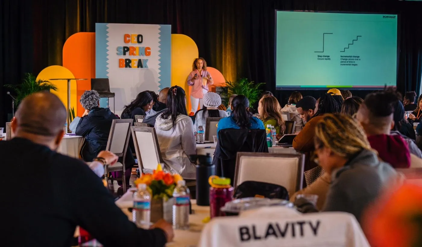 Attendees at a conference session watching a speaker on a stage with a branded backdrop