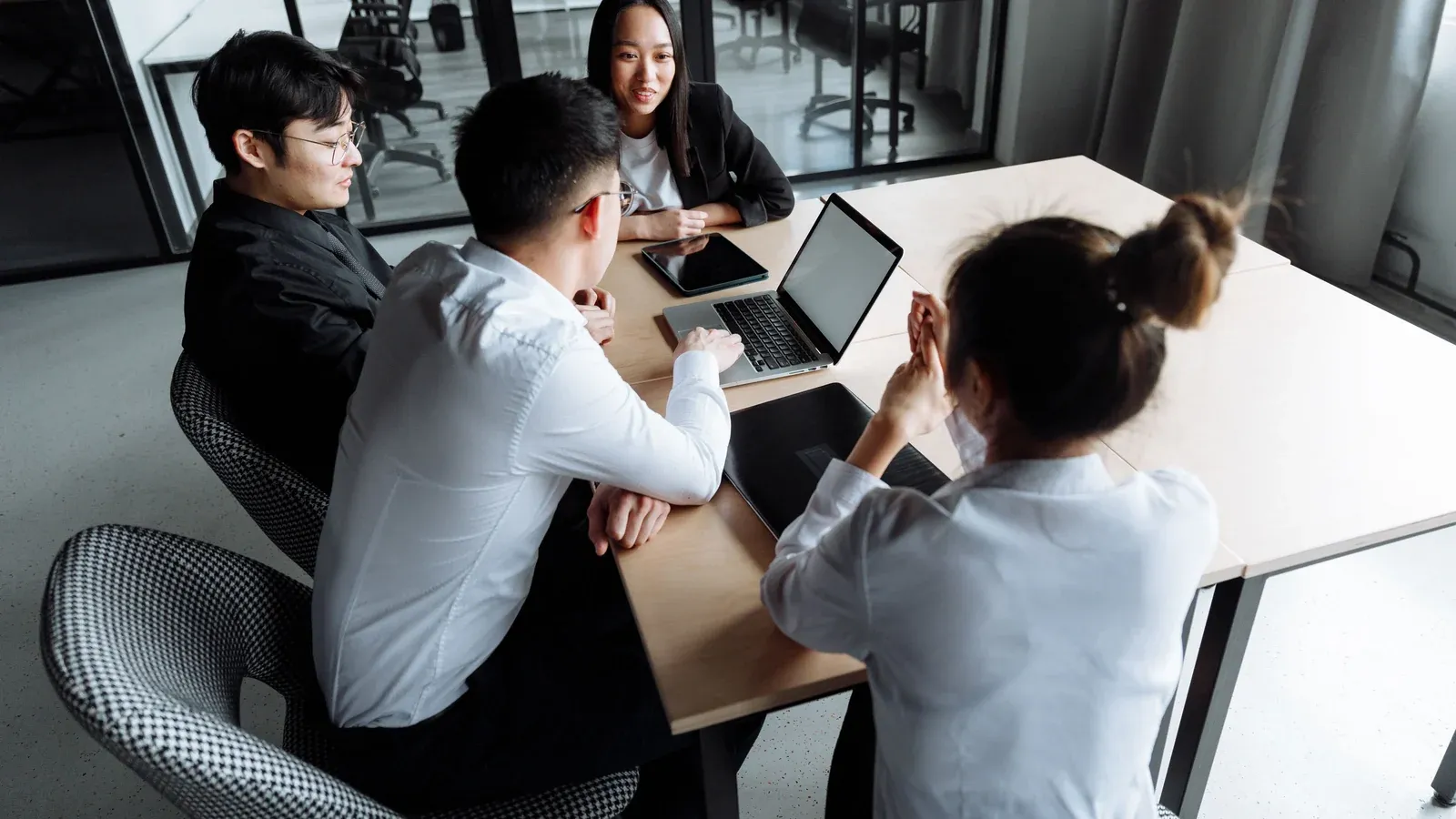 Four colleagues gather around a corner table with a laptop and tablet in a glass-walled meeting room during a daytime session at a company offsite.