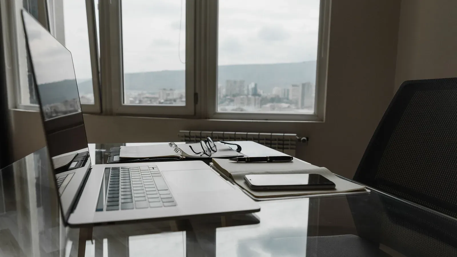 A laptop, notebook, pen and phone rest on a glass desk beside tall windows with a hazy city view.