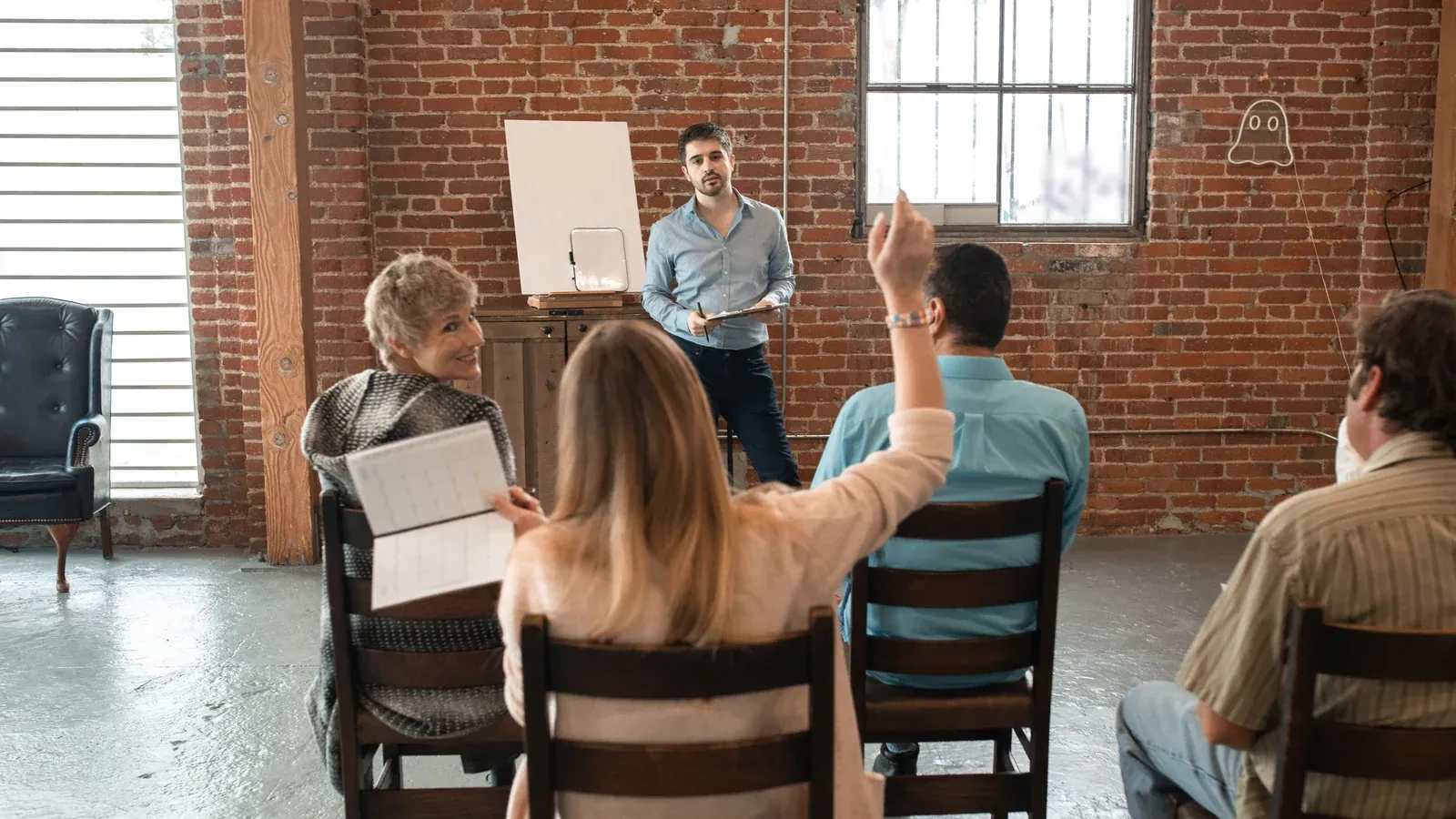 A facilitator stands beside a blank flip chart in a brick-walled loft as four seated participants listen and one raises a hand.