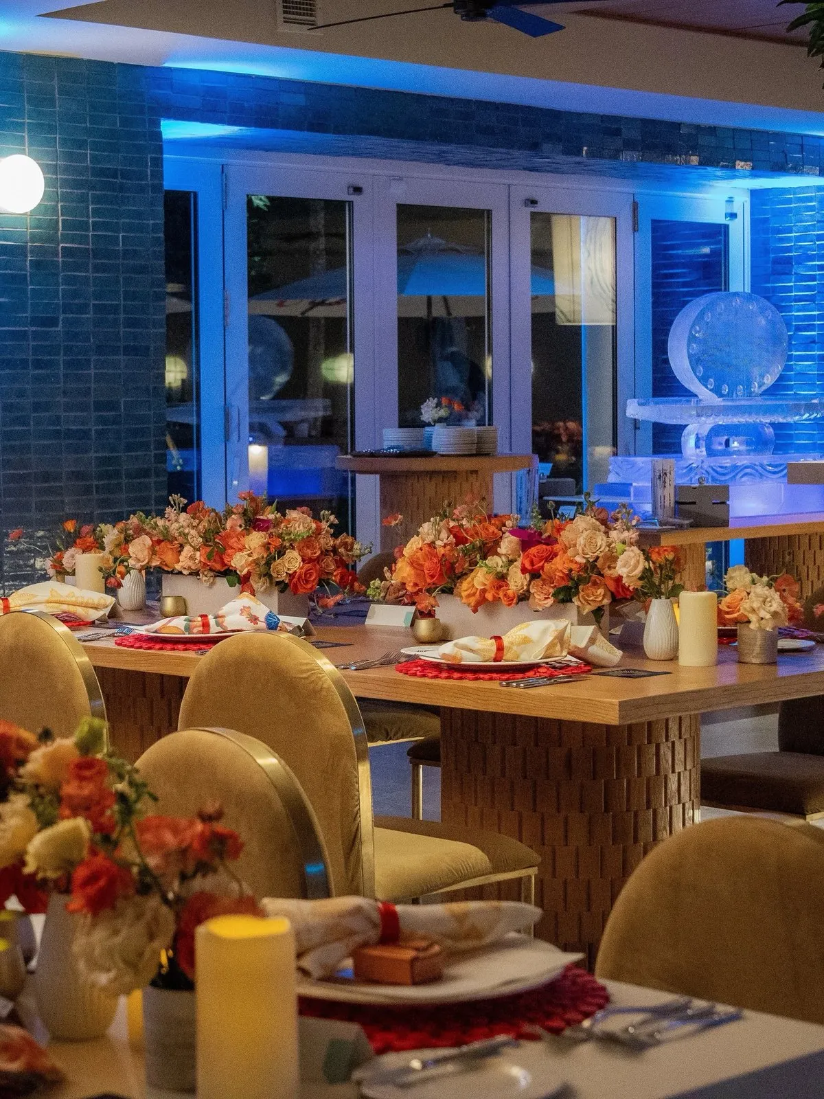 Dinner table set with floral centerpieces and gold chairs beside blue-lit windows