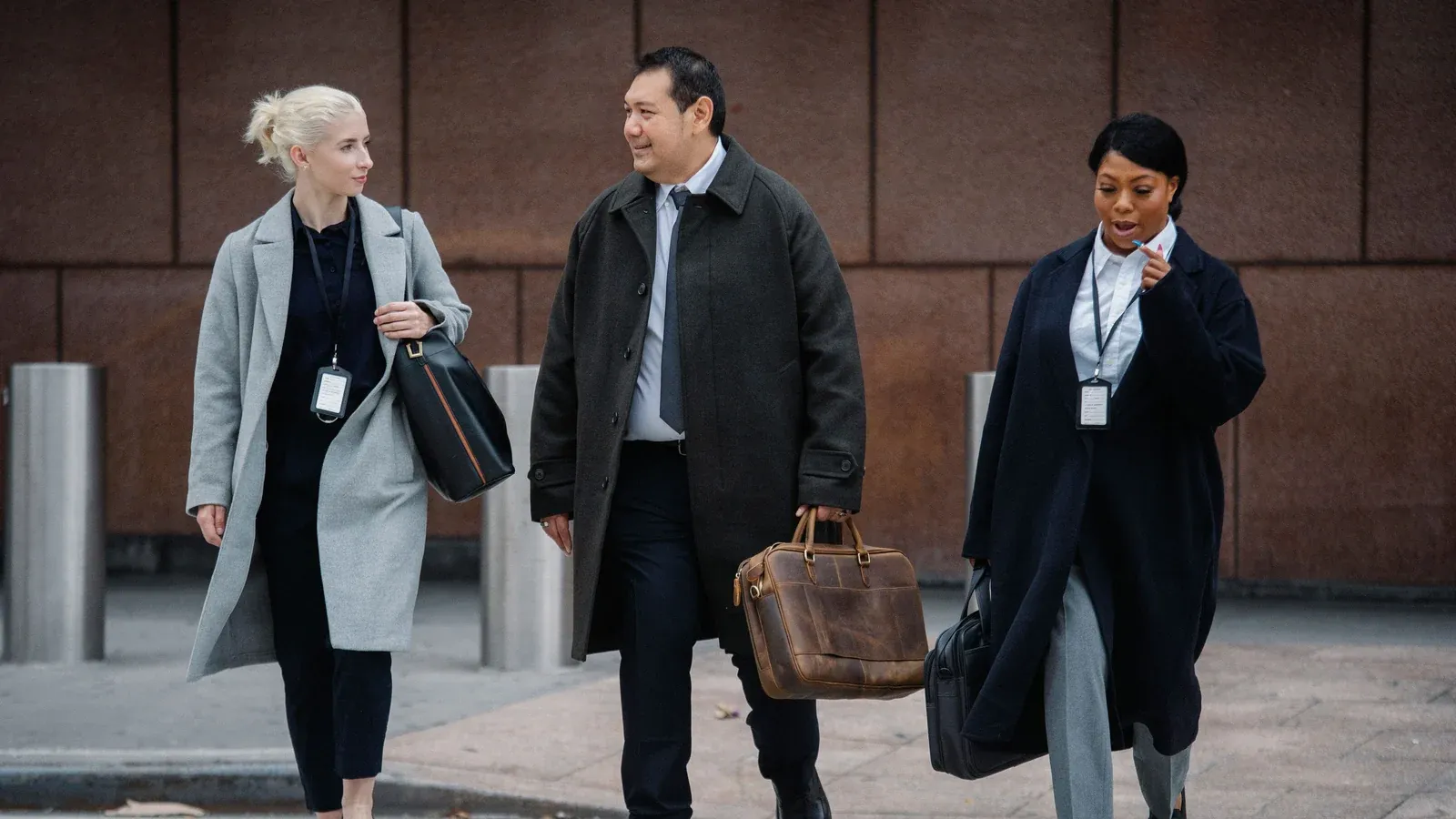 Three colleagues in long coats walk together outside an office building. They carry work bags and talk as they head to their next session.