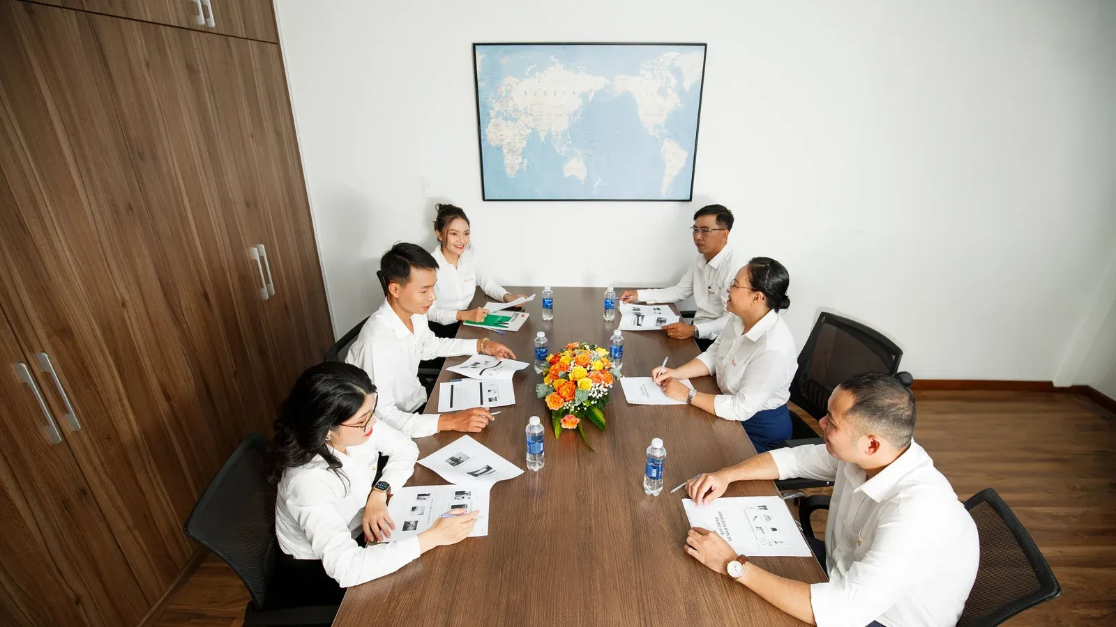 A small team sits around a long wooden table in a bright meeting room. Papers, water bottles and a vase of flowers sit between them as they talk.