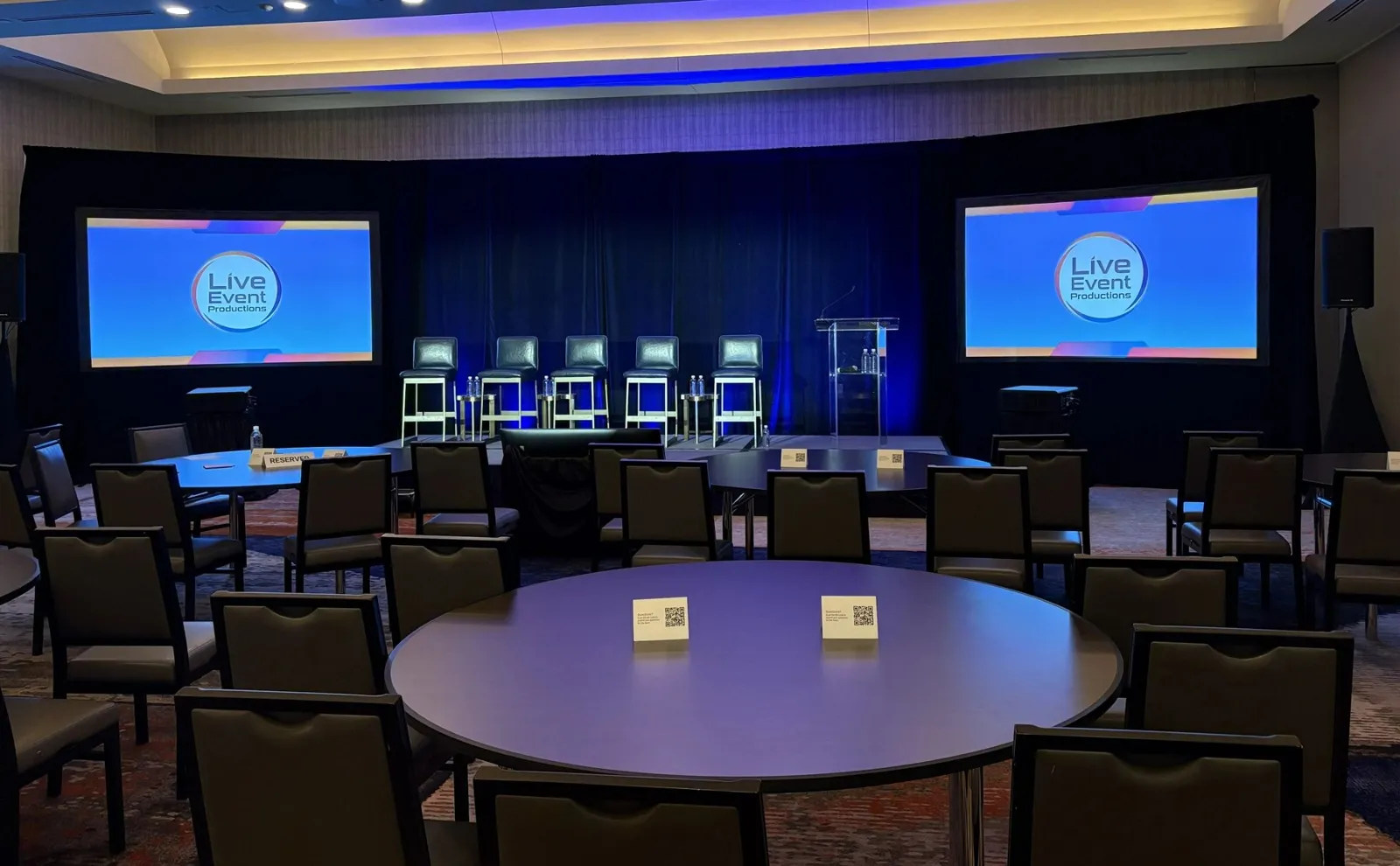 Ballroom with a stage, side screens and round tables set for a corporate program