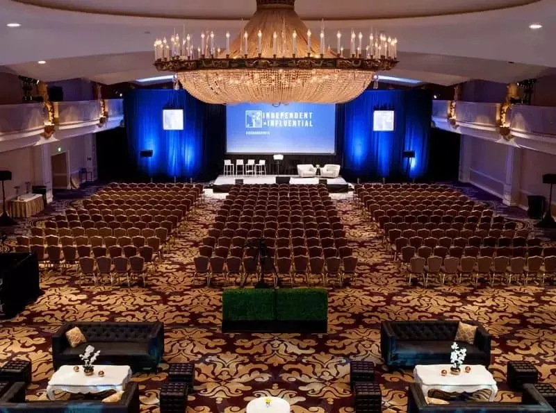Theater-style seating facing a stage with screens in a chandeliered hotel meeting room