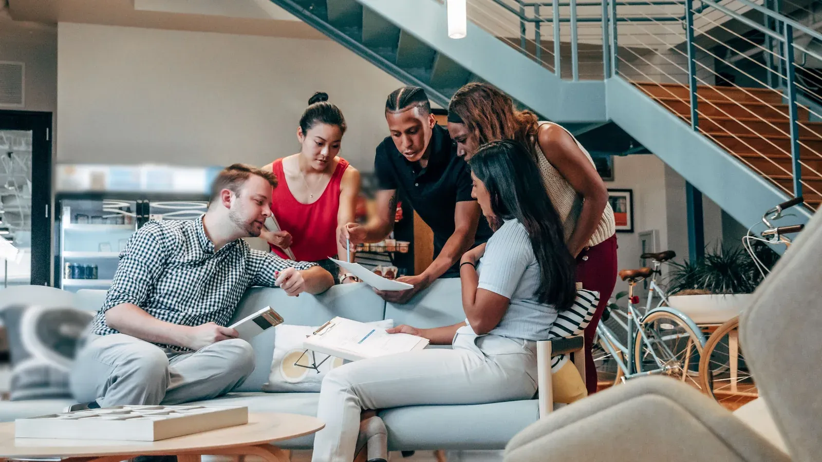Five colleagues in casual clothes gather around a sofa in an office lobby, reading a sheet of paper.