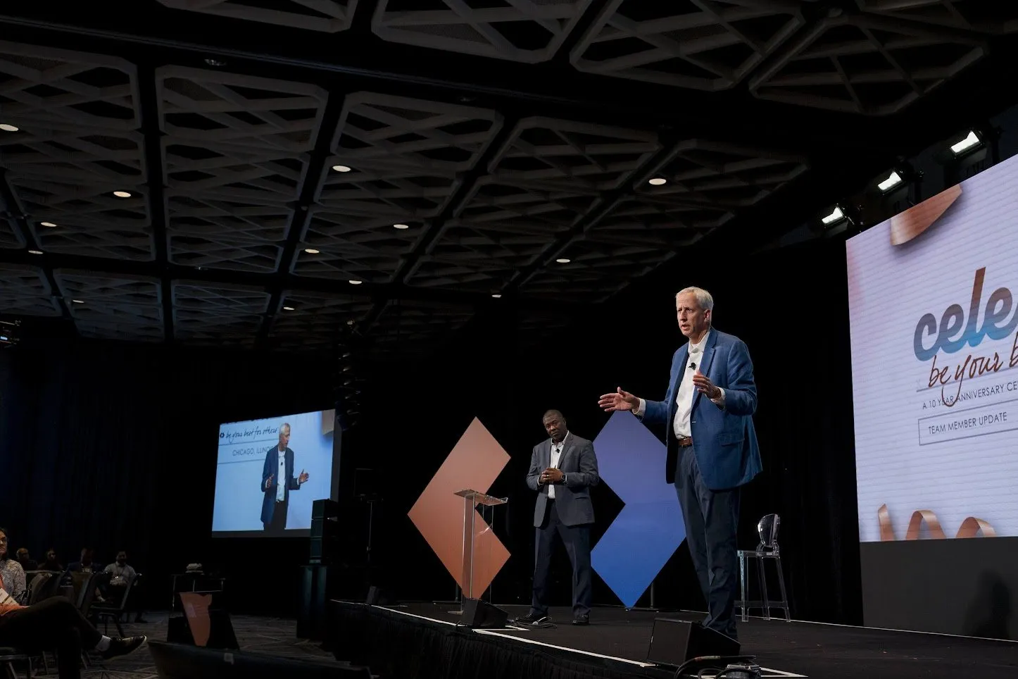 Speaker on a dark stage with screens during a company update