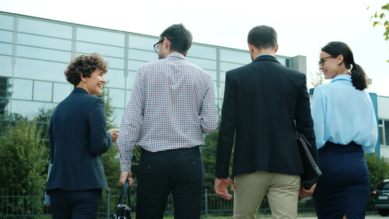 Four colleagues in smart business dress walk together toward a glass office building, chatting as they go. Green shrubs and fresh leaves line the path on a bright spring day.