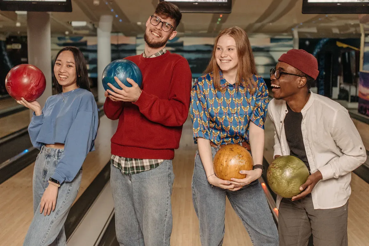 Four colleagues line up along a bowling lane, each holding a ball and laughing as they wait for their turn. An evening at the lanes gives the team something to do together beyond the usual drinks.