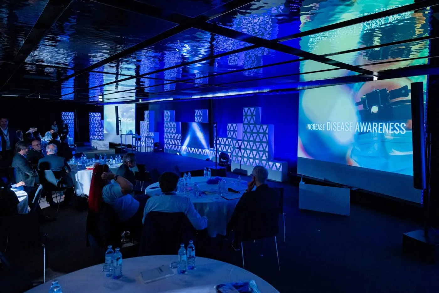 Dark ballroom with round tables and large projected screens at an evening program