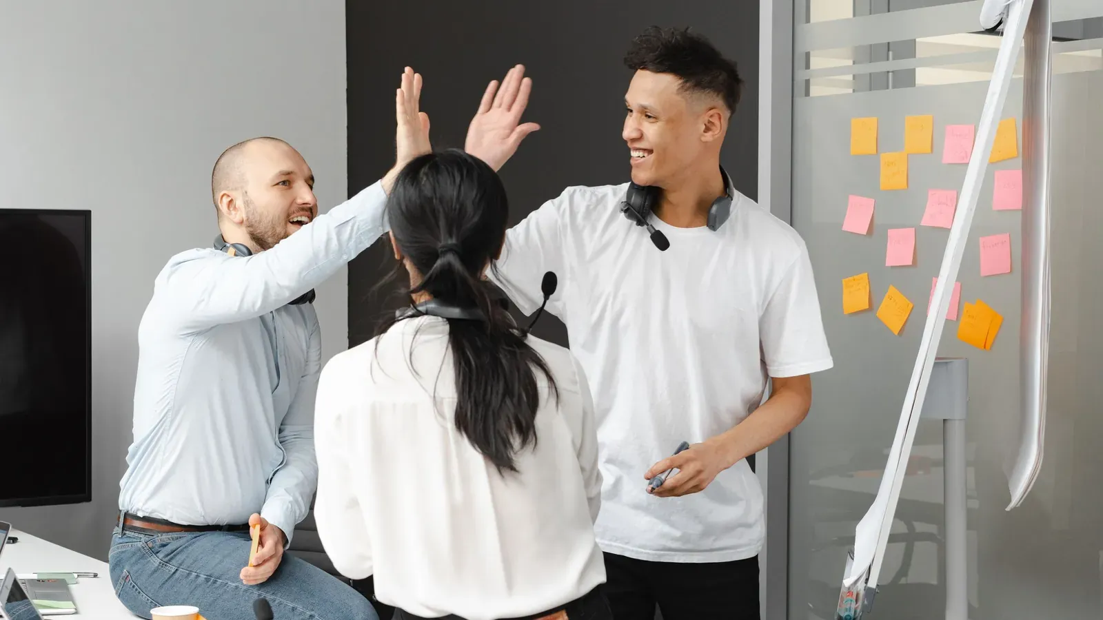 Three coworkers wearing headsets around their necks share a double high five beside a wall of blank sticky notes.