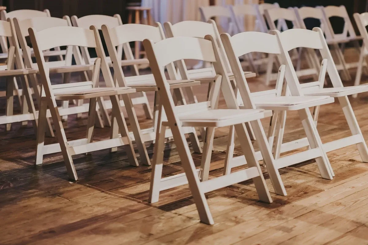 Rows of white wooden folding chairs stand on a worn hardwood floor in an event venue.