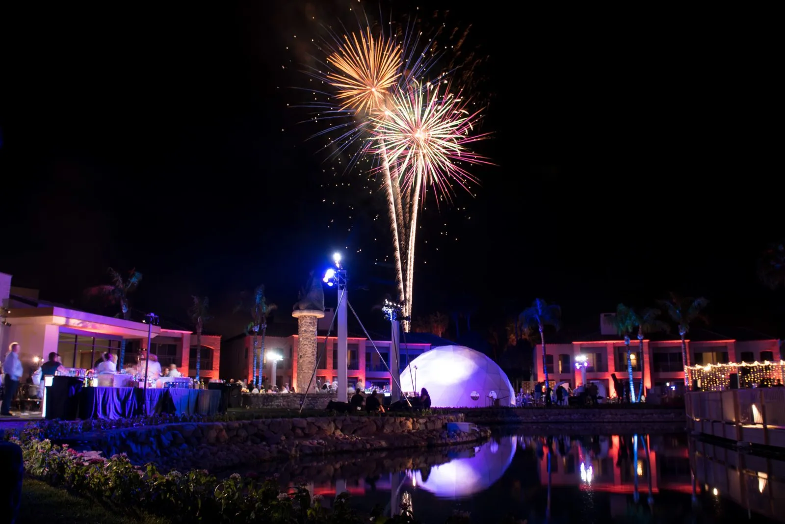 Fireworks over a lit outdoor reception with a domed structure and string lights at night