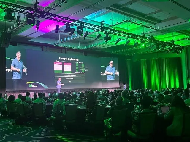 Presenters on stage under green and purple lighting in front of a seated audience