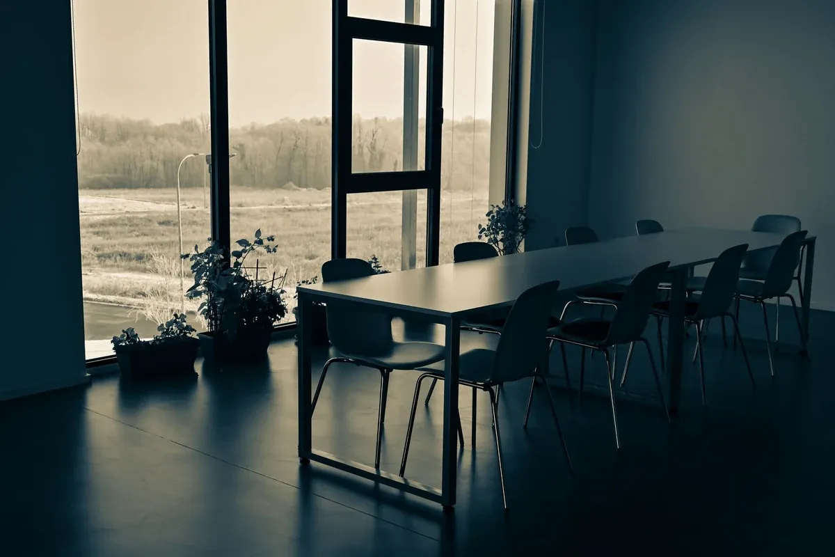 A long white meeting table with simple shell chairs sits in a quiet, dim room beside floor-to-ceiling windows. Potted plants line the window ledge, and a hazy field and bare trees are visible outside.