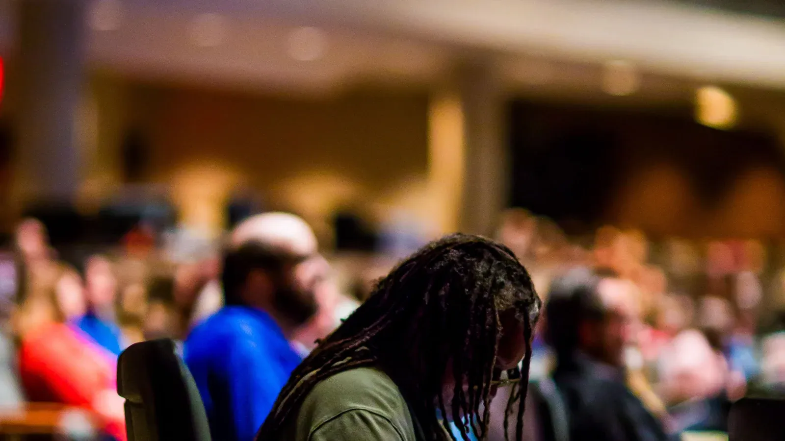 Attendees sit in rows and listen during a session in a large conference hall. Warm lights glow over the softly blurred crowd.
