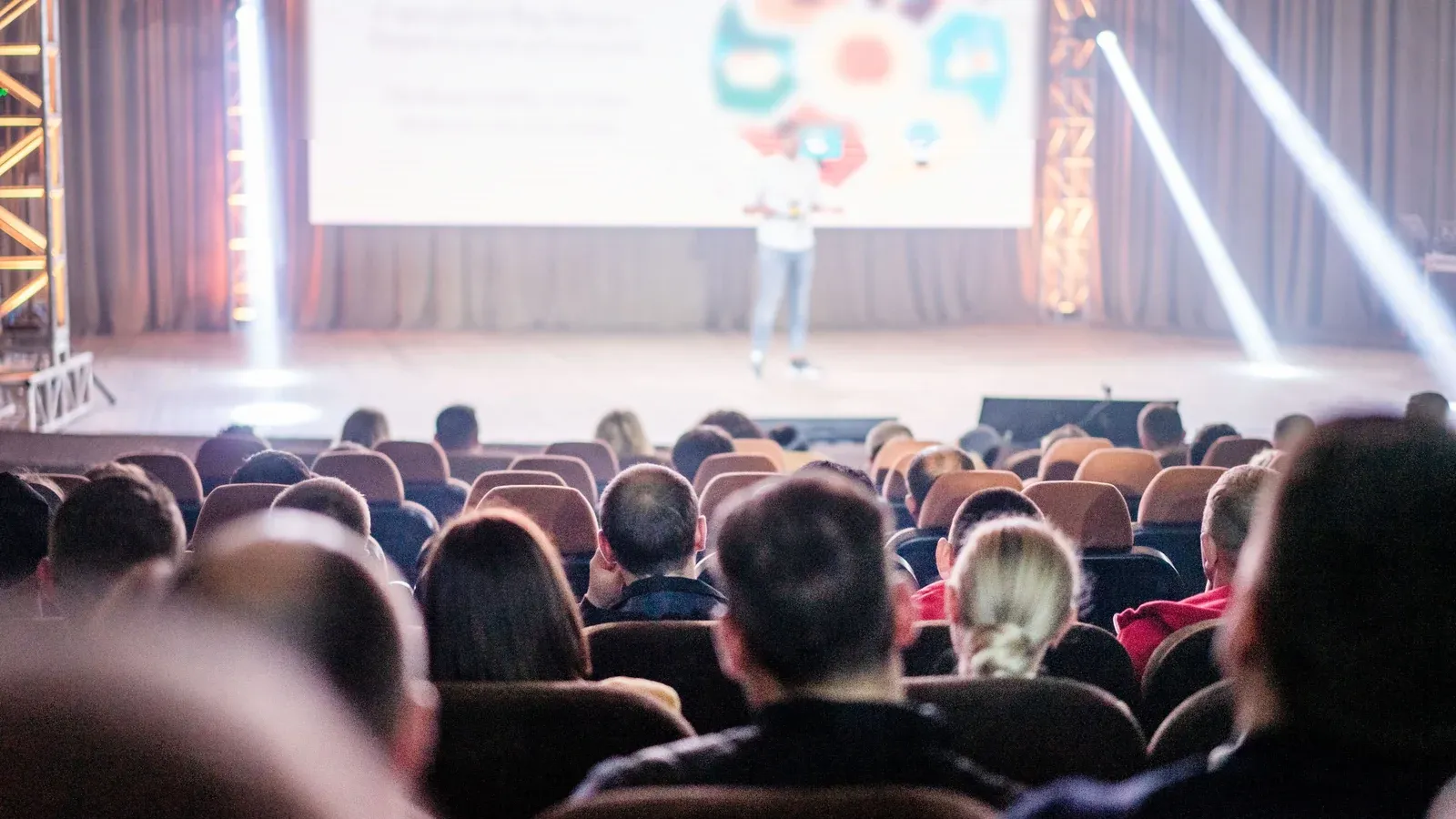 A full audience sits in rows of padded seats, facing a presenter on a wide stage with a large screen behind him. Soft beams of stage light cut across the hall as the session gets under way.