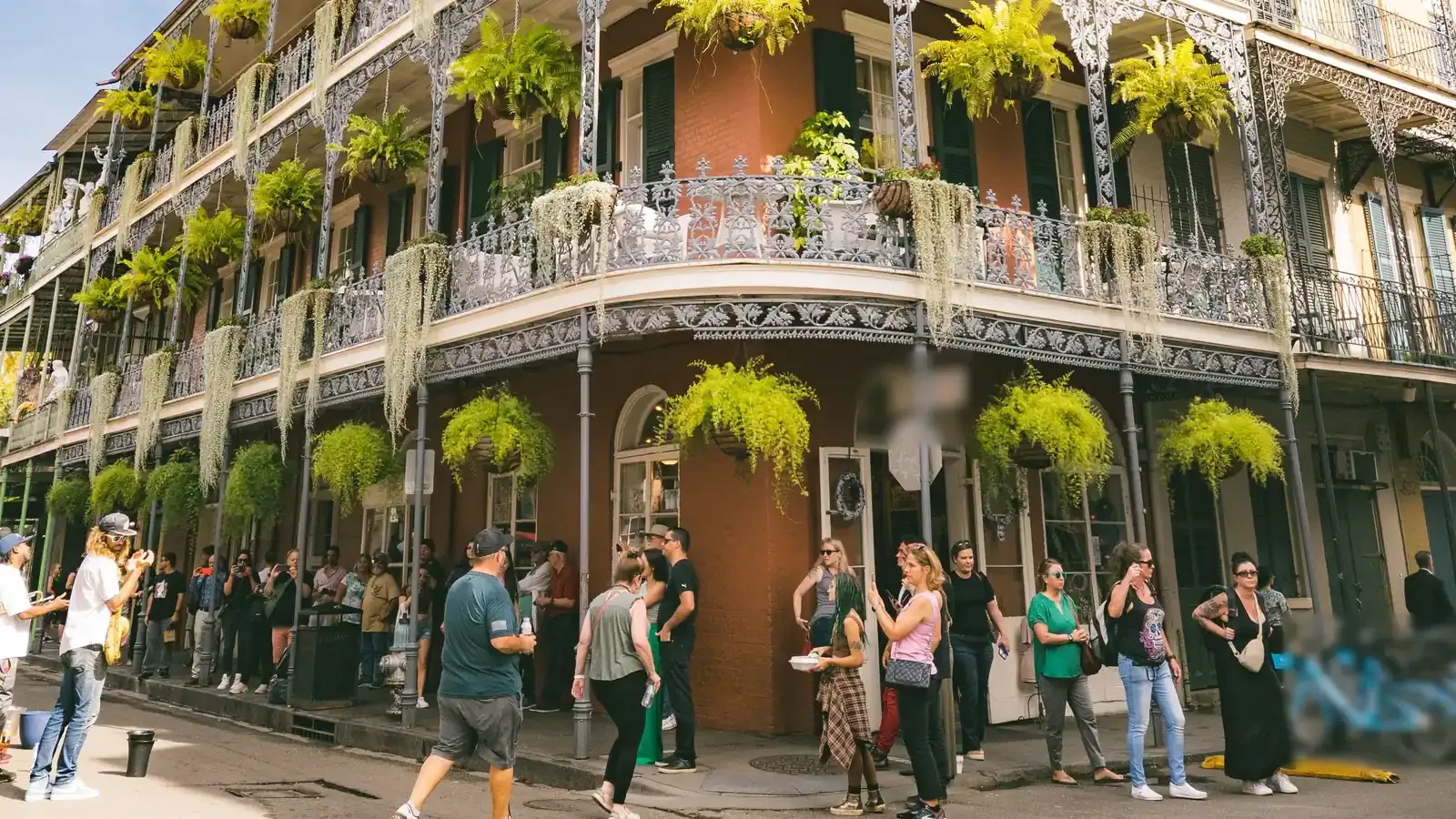 Visitors stroll past a corner building wrapped in lacy iron balconies and hanging ferns on a sunny day during an incentive trip in New Orleans.