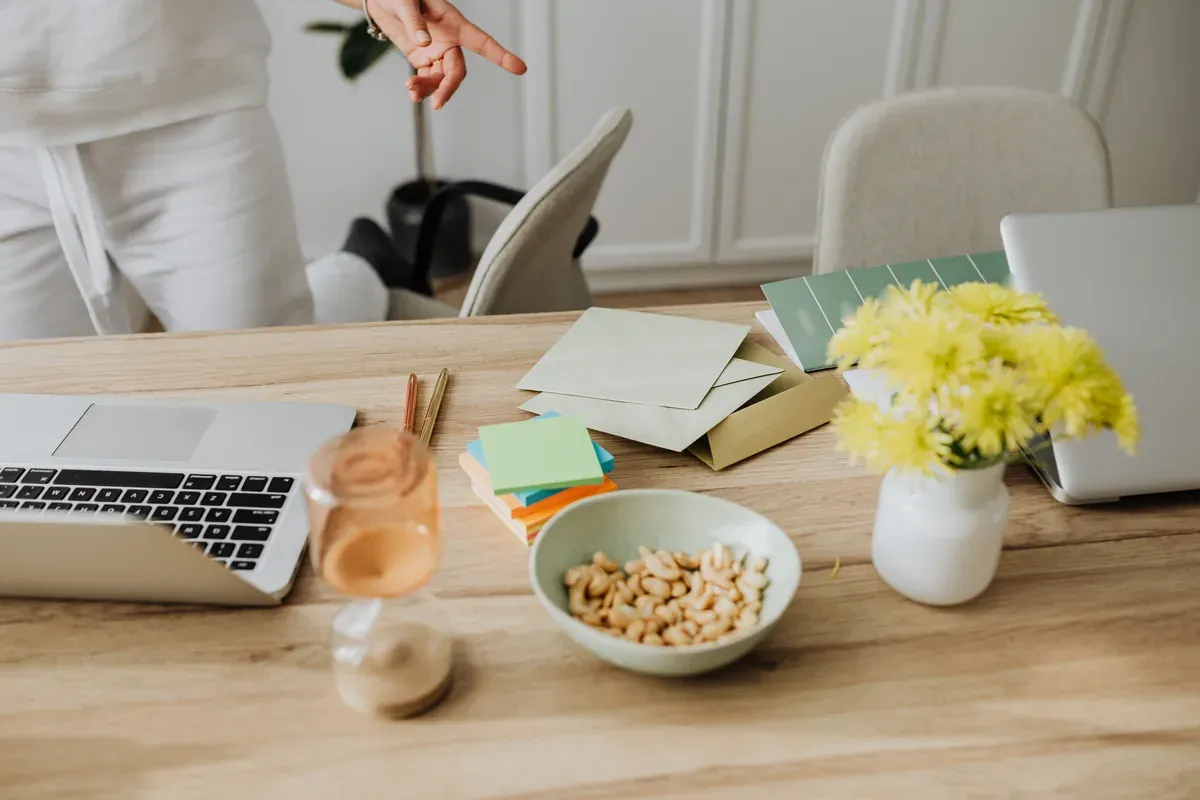 An open kit of cards and envelopes sits on a light wooden home desk between two laptops, with a bowl of cashews, sticky notes and fresh yellow flowers. It suggests a remote team member settling in to join the celebration from home.