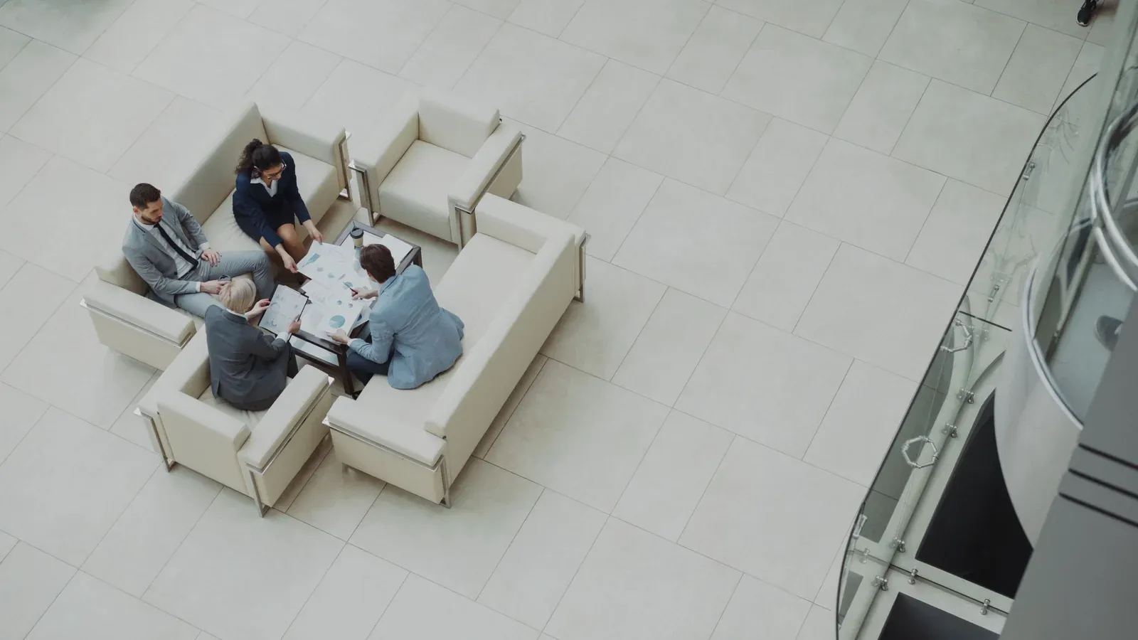 Seen from a balcony above, four colleagues review charts around a low table in a bright lobby lounge during a working session at a company offsite.