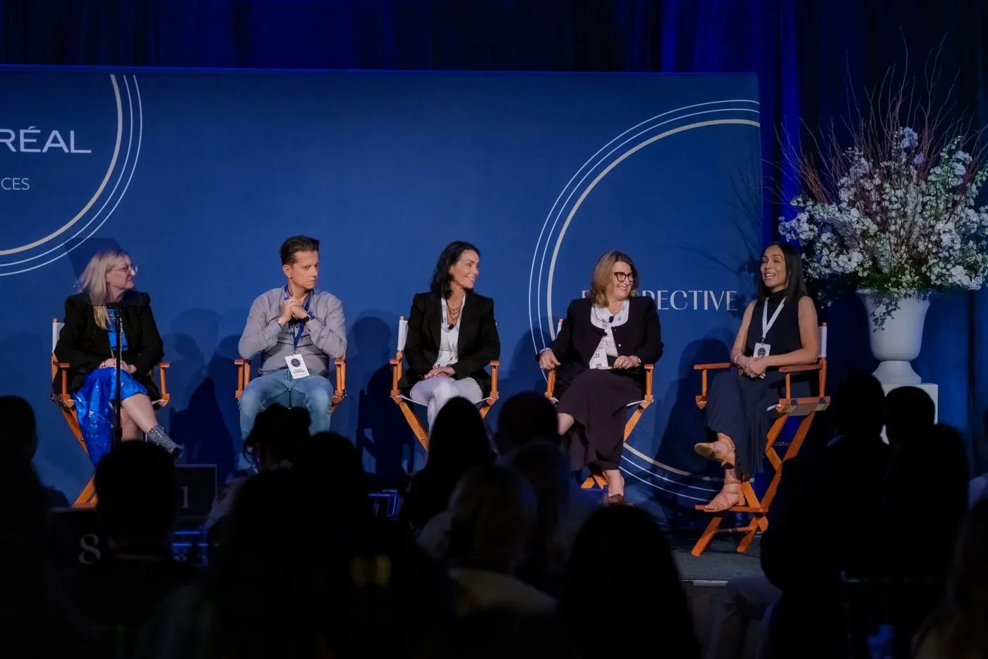 Panel of five speakers seated in front of a blue backdrop with a floral arrangement