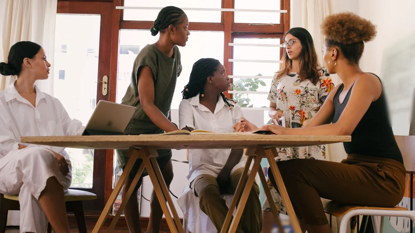 Five colleagues talk around a wooden trestle table with a notebook and a laptop beside tall sunlit windows during a company offsite.