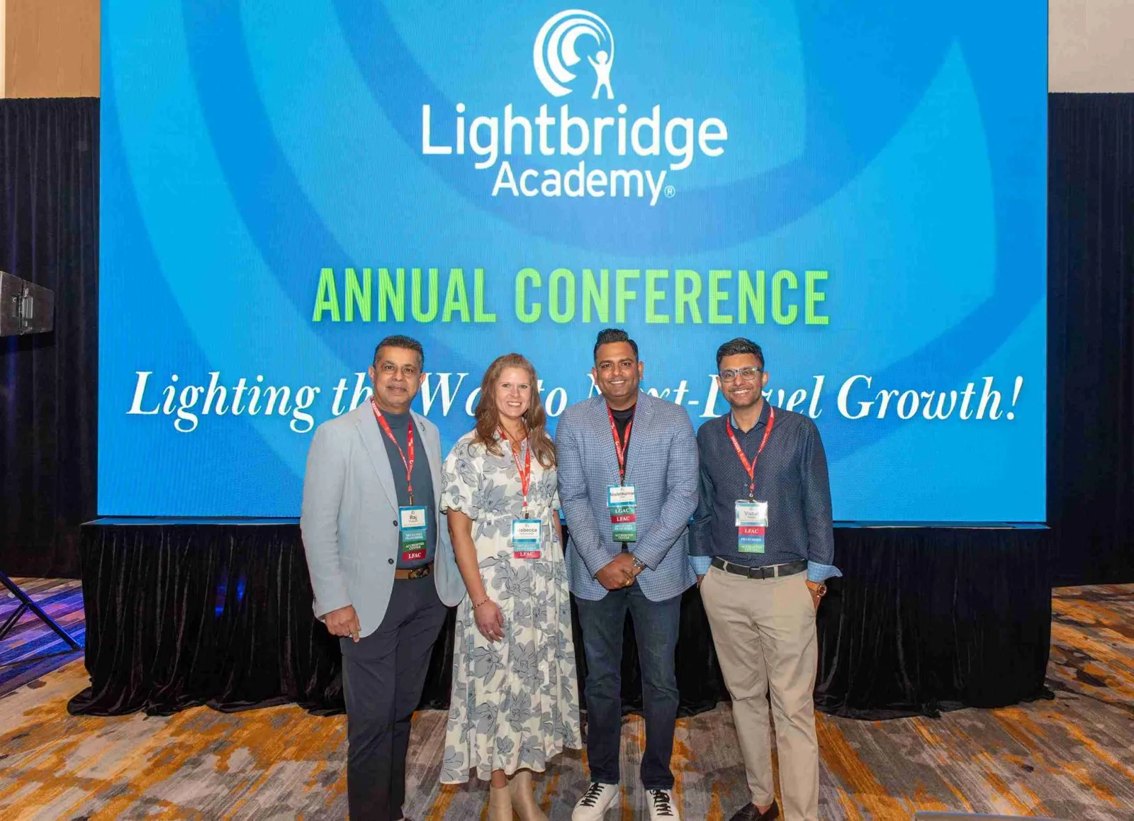 Four attendees in front of a Lightbridge Academy annual conference screen