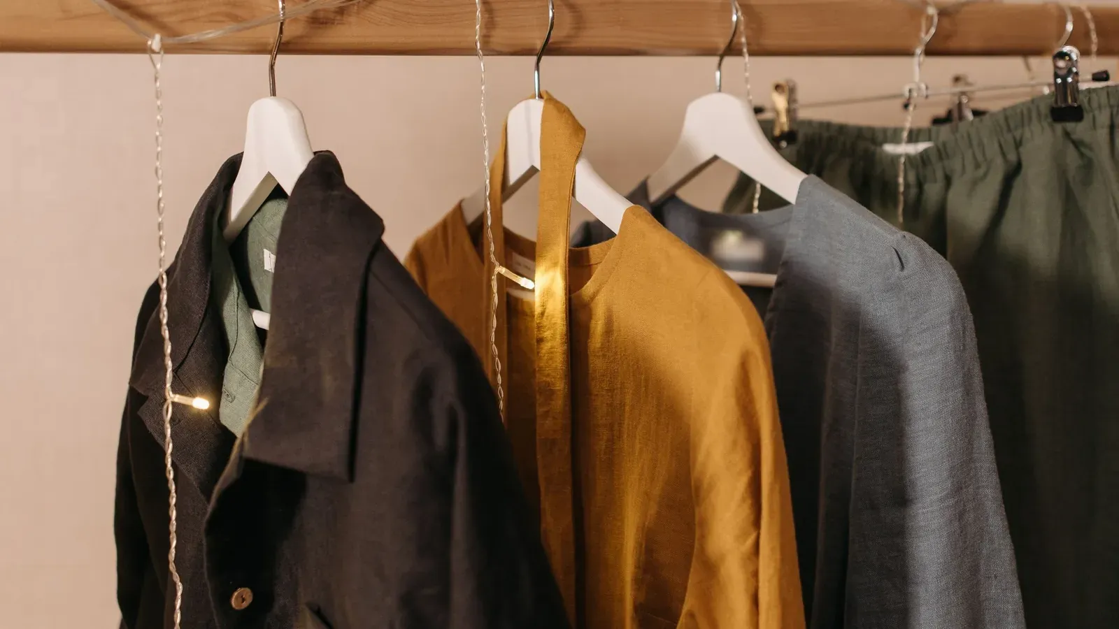 Black, mustard, and gray linen jackets hang on white hangers from a wooden rail strung with small fairy lights.