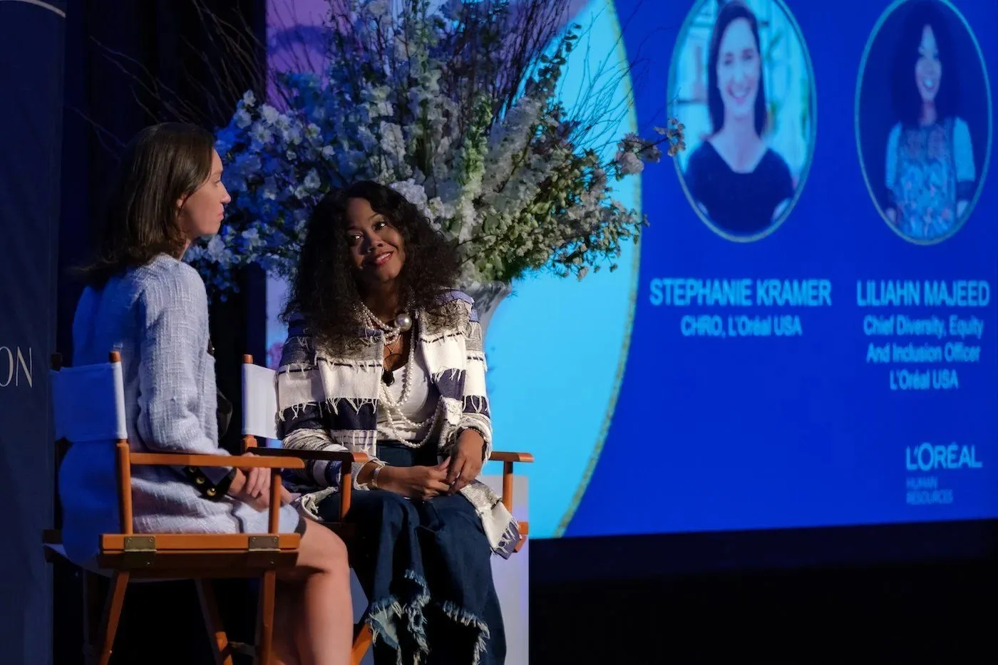 Two speakers in a fireside conversation on a stage with a large branded screen