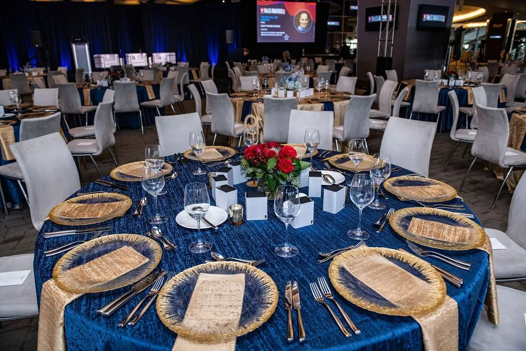 Round dinner tables with gold chargers and blue linens in a hotel ballroom