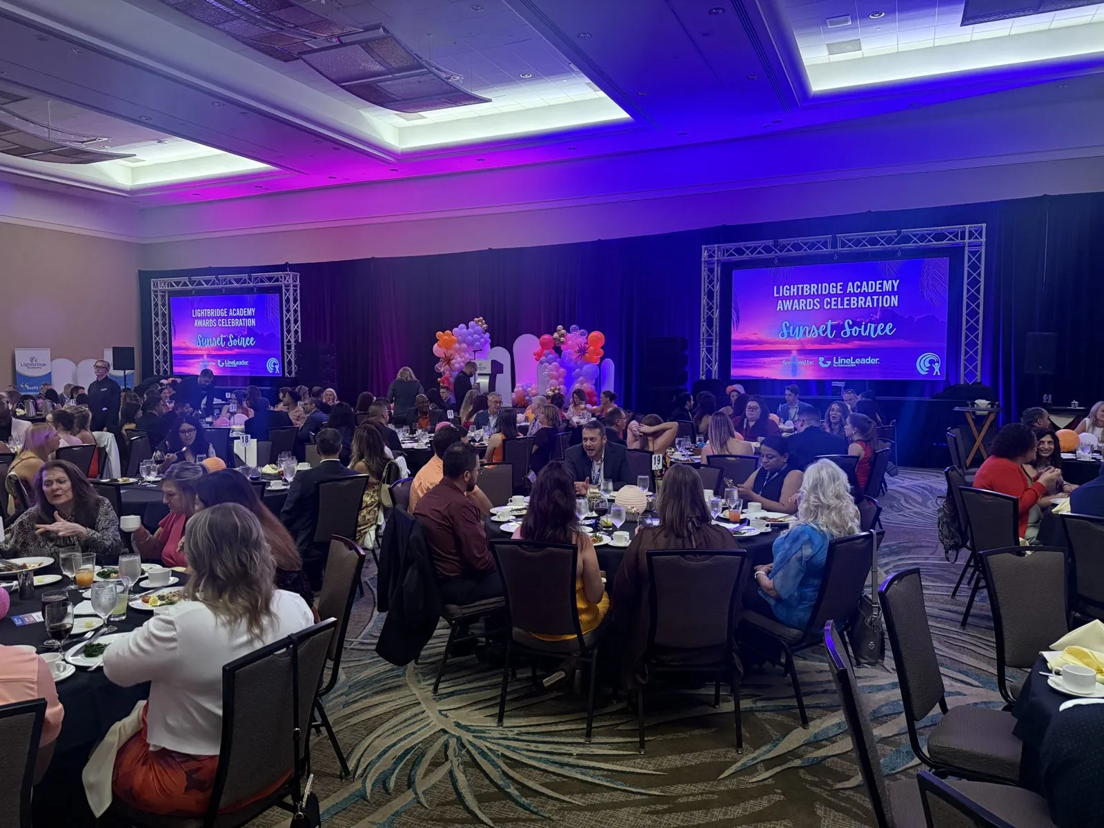 Ballroom of seated guests with purple lighting and a stage screen