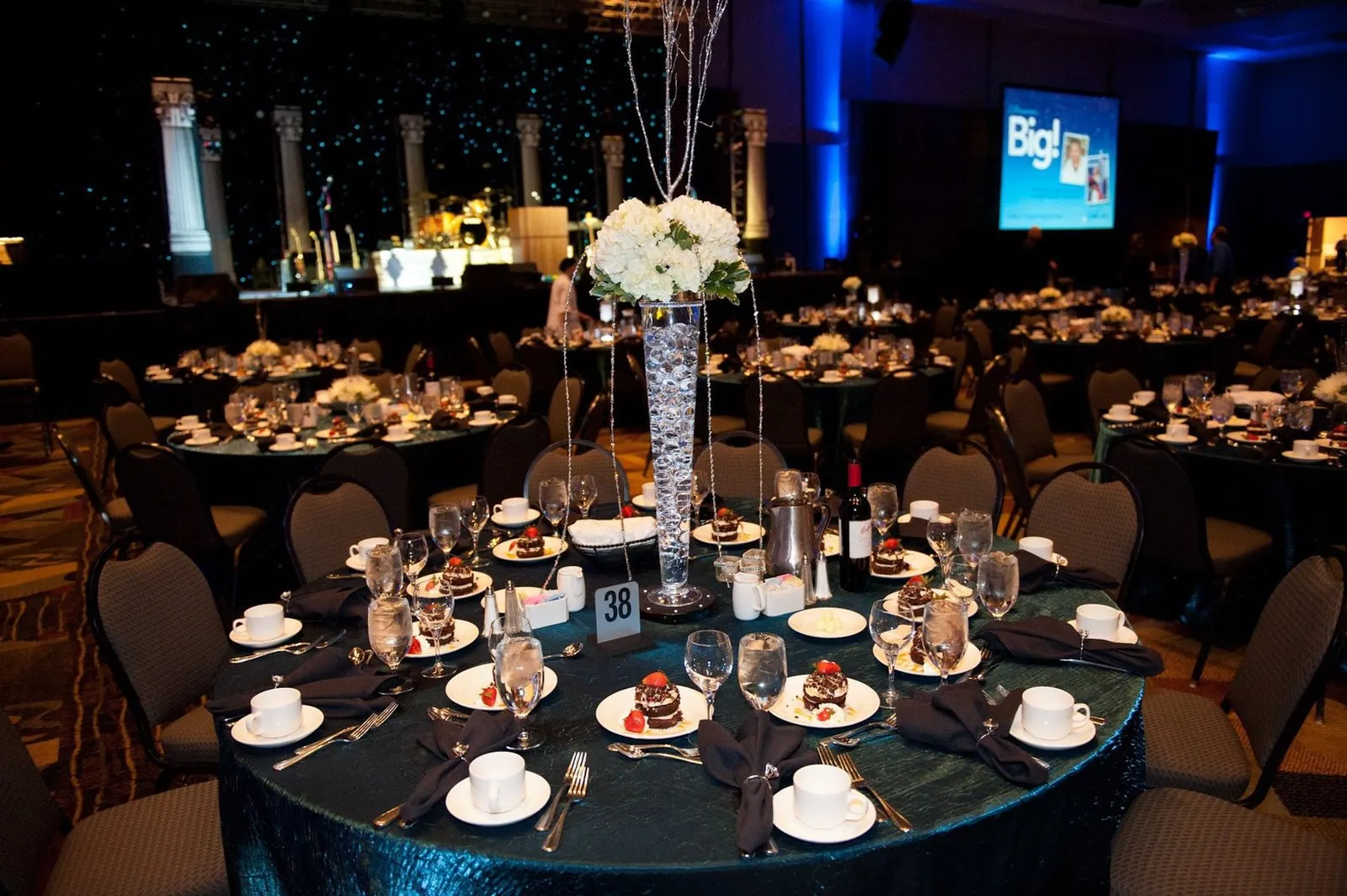 Ballroom gala dinner with blue uplighting, tall centerpieces and stage screens