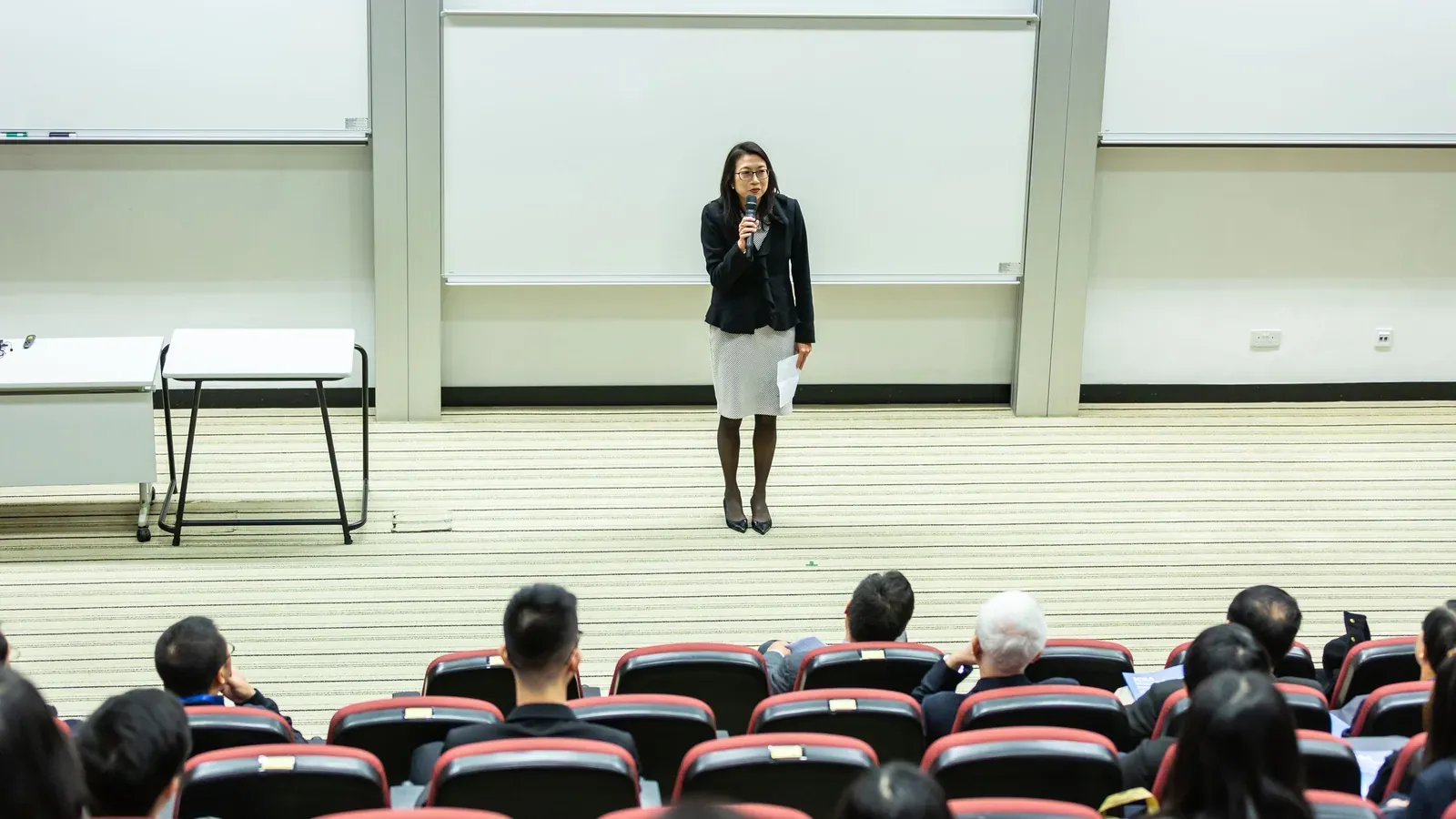 A speaker in a dark blazer holds a microphone on a bare stage while seated attendees face them in red auditorium chairs.
