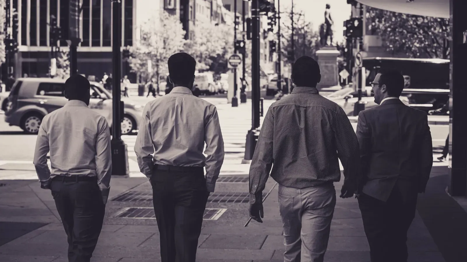 Four colleagues in business clothes walk away from the camera along a city sidewalk in a black and white photo.
